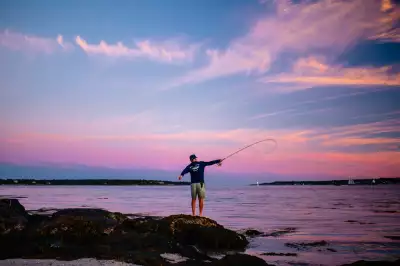 An angler fishes at sunset from Diamond Island Ledge in Casco Bay, framed by a wash of pink and blue sky as evening settles over the water