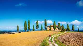 Classic panorama view of scenic Tuscany landscape with farmhouse and male cyclist on a country road on a beautiful sunny day in summer, Italy