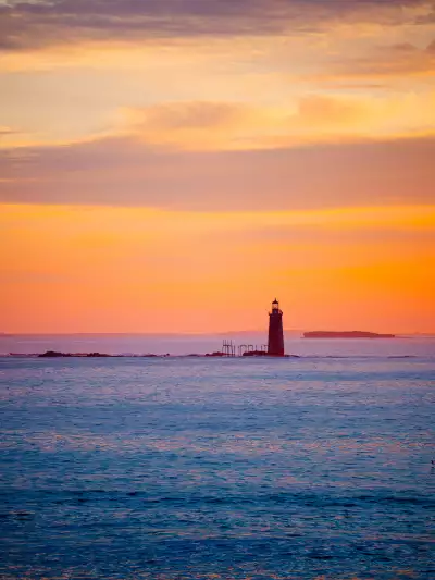 Ram Island Ledge Light stands silhouetted against a fiery sunset in Casco Bay, its granite tower marking the approach to Portland